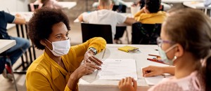 Black teacher with a face mask explaining exam results to elementary student in the classroom.